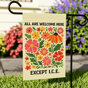 May include: A cream garden flag with the text "ALL ARE WELCOME HERE EXCEPT I.C.E." is displayed outdoors. The flag features a colourful floral design with red, pink, orange, and yellow flowers. The flag is hanging on a black metal stand in a grassy garden.