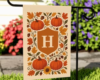 Könnte beinhalten: Eine beige Gartenflagge mit Herbstmotiv. Die Flagge zeigt vier orangefarbene Kürbisse, Herbstblätter und den Buchstaben "H" in einem Schild. Eine Sonnenblume und Eicheln sind ebenfalls enthalten. Die Flagge wird auf einem schwarzen Metallständer präsentiert.