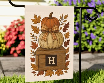Könnte beinhalten: Eine cremefarbene Gartenflagge mit einem herbstlichen Design. Das Design zeigt gestapelte Kürbisse, eine karierte Schleife und Herbstblätter. Eine Holzkiste mit dem Buchstaben "H" befindet sich am Fuß der Kürbisse. Die Flagge wird auf einem schwarzen Metallständer angezeigt.
