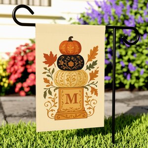 Peut inclure: Un drapeau de jardin beige présente un motif de citrouilles empilées en orange, noir et crème, avec des feuilles d'automne et la lettre "M". Le drapeau est sur un support en métal noir dans un jardin herbeux.