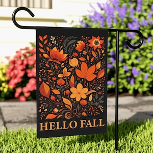以下が含まれることがあります： 「HELLO FALL」の文字が金色で書かれた黒いガーデンフラッグ。オレンジ、ゴールド、ブラウンの色合いの花柄のデザインが特徴です。芝生の庭にある黒い金属製のスタンドに飾られています。
