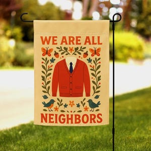 May include: A beige garden flag displaying the text "WE ARE ALL NEIGHBOURS" in orange and red. The flag features a red cardigan, a white shirt, and a blue tie design. Decorative elements include butterflies, birds, and floral patterns.