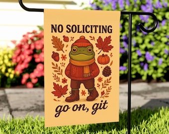 Könnte beinhalten: Eine beige Gartenflagge mit einer Cartoon-Fröschin in rotem Pullover, Mütze und Schal, umgeben von Herbstblättern und einem Kürbis. Die Flagge trägt die Aufschrift "NO SOLICITING" und "go on, git". Sie wird auf einem schwarzen Metallständer im Garten gezeigt.