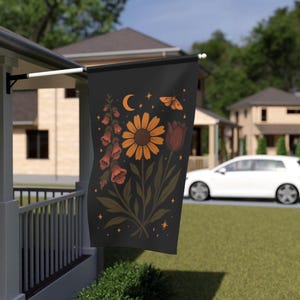 May include: A black garden flag featuring a floral design with a sunflower, a tulip, and other flowers. The flag also includes a crescent moon, a butterfly, and stars. The flag is hanging on a white pole on a porch.