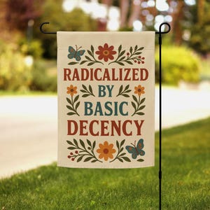 May include: A beige garden flag with the text "RADICALIZED BY BASIC DECENCY" in a mix of colors. The flag is decorated with floral and butterfly designs in shades of orange, brown, and teal. The flag is hanging on a black metal stand in a grassy yard.