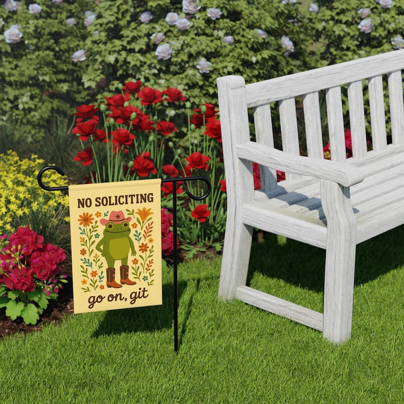 May include: A small garden flag with the text "NO SOLICITING" and a cartoon frog wearing a cowboy hat and boots. The flag is on a black metal stand, set in a grassy yard with a white bench and colorful flowers in the background.