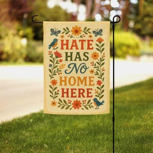 May include: A cream-coloured garden flag with the words "HATE HAS NO HOME HERE" in red, blue, and orange. The flag has floral and butterfly designs. A black metal stand holds the flag in a grassy garden.