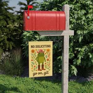 May include: A red mailbox with a small red flag, mounted on a wooden post. A decorative garden flag hangs below, featuring a cartoon frog in a cowboy hat and boots, with the words "NO SOLICITING" and "go on, git".