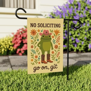 May include: A beige garden flag with a cartoon frog wearing a cowboy hat and boots, surrounded by flowers. The flag reads "NO SOLICITING" at the top and "go on, git" at the bottom. The flag is displayed on a black metal stand in a grassy yard.
