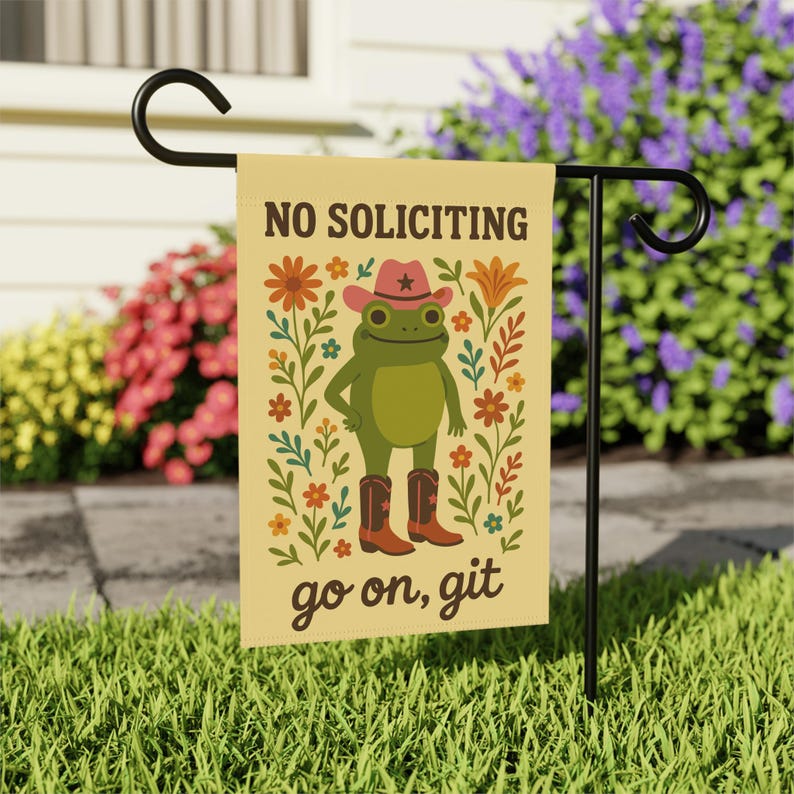 May include: A beige garden flag with a cartoon frog wearing a cowboy hat and boots, surrounded by flowers. The flag reads "NO SOLICITING" at the top and "go on, git" at the bottom. The flag is displayed on a black metal stand in a grassy yard.