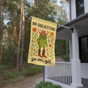 May include: A yellow house flag with the text "NO SOLICITING" and "go on, git". The flag features a cartoon frog wearing a cowboy hat and boots, surrounded by floral designs. The flag is hanging on a porch.