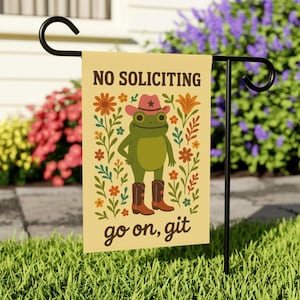 Puede incluir: Una bandera de jardín con una rana con sombrero y botas de vaquero, rodeada de flores. La bandera dice "NO SOLICITING" y "go on, git". La bandera está en un soporte de metal negro en un jardín.
