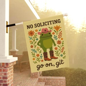 May include: A rectangular, cream-colored garden flag with the text "NO SOLICITING" and "go on, git". The flag features a cartoon frog wearing a pink cowboy hat and boots, surrounded by floral illustrations.