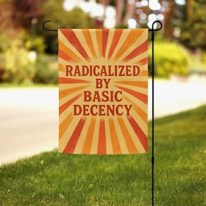 Może przedstawiać: Flaga ogrodowa z napisem "RADICALIZED BY BASIC DECENCY" w stylu retro. Flaga ma promienisty wzór w odcieniach pomarańczu i beżu. Jest umieszczona na czarnym metalowym słupku w trawiastym ogrodzie.