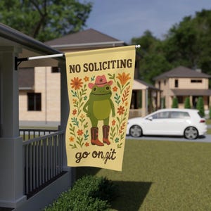 May include: A rectangular, cream-colored flag with the text "NO SOLICITING" at the top. The flag features a cartoon frog wearing a cowboy hat and boots, surrounded by floral designs. The words "go on, git" are at the bottom.
