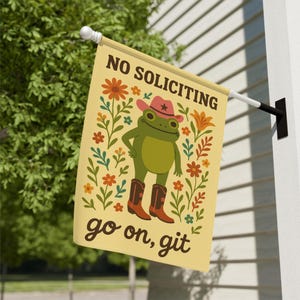 May include: A rectangular, cream-colored flag with the words "NO SOLICITING" and "go on, git" in brown text. The flag features a cartoon frog wearing a cowboy hat and boots, surrounded by floral illustrations.