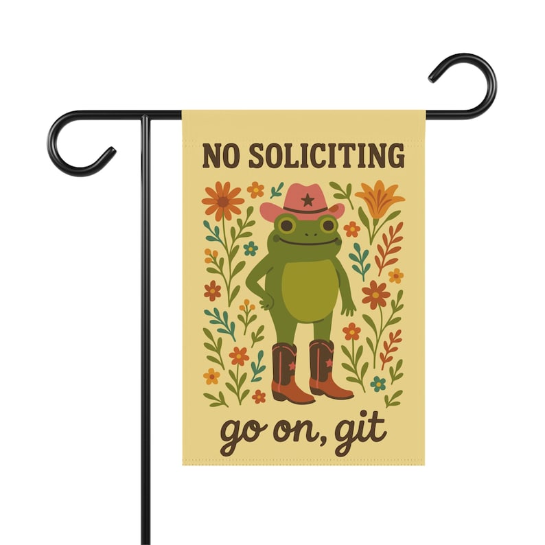 May include: A garden flag with a cartoon frog wearing a cowboy hat and boots, surrounded by flowers. The flag reads "NO SOLICITING" and "go on, git" in a playful font. The flag is attached to a black metal stand.