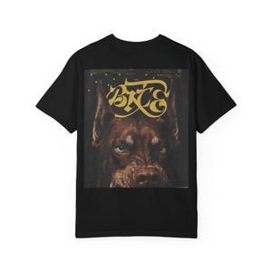 Op de afbeelding: Zwart T-shirt met een close-up van een bruine hond met blauwe ogen en het woord "BITE" in gouden letters. De hond heeft een serieuze uitdrukking. Het shirt is een klassieke ronde hals.