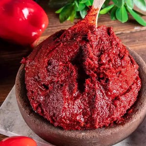 Op de afbeelding: Een close-up van een kom gevuld met een dikke, dieprode pasta, mogelijk tomatenpuree of een soortgelijk condiment. Een houten lepel is gedeeltelijk in de pasta gestoken. Rode paprika's en groene bladeren zijn zichtbaar op de achtergrond.