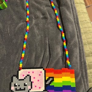 Puede incluir: Un collar de cuentas de colores con un diseño pixelado de Nyan Cat. El gato es gris con un cuerpo rosa y una estela de arcoíris. La correa del collar está hecha de cuentas de colores del arcoíris.