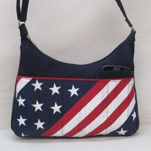 May include: A navy blue shoulder bag with a patriotic design. The front features a pattern of white stars on a blue background and red and white stripes. The bag has a pocket and a black strap.