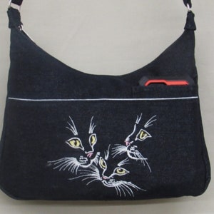 May include: Black denim shoulder bag with a white embroidered design of three cat faces. The bag has a small pocket on the front with a red phone case inside.