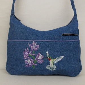 May include: Blue denim shoulder bag with a hummingbird and flower embroidery. The bag has a front pocket, a purple trim, and a zipper closure. The hummingbird is green, red, and white, and the flowers are purple.