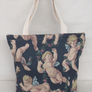May include: A tote bag with a dark blue background featuring a repeating pattern of cherubs with wings and floral garlands. The bag has cream-colored handles and a zipper closure. The bag is made of a woven fabric.