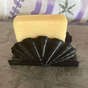 May include: A rectangular bar of light yellow soap rests in a black, fan-shaped soap dish. The soap dish has a textured, shell-like design. The background is a patterned fabric with purple and green elements.