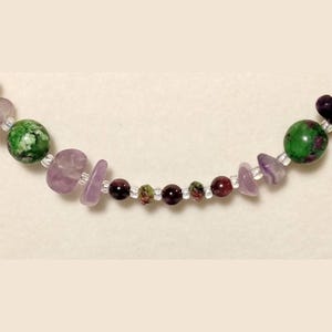 May include: A multi-colored beaded necklace with green, purple, and red beads. The necklace is made with small, round beads and larger, irregularly shaped beads.