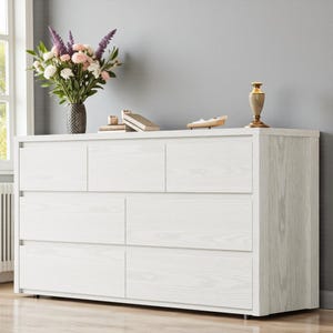 Puede incluir: Una c&oacute;moda de madera blanca con seis cajones y un acabado de veta de madera clara. La c&oacute;moda est&aacute; coronada con un jarr&oacute;n de flores, libros y objetos decorativos. La habitaci&oacute;n tiene una pared gris y una ventana.