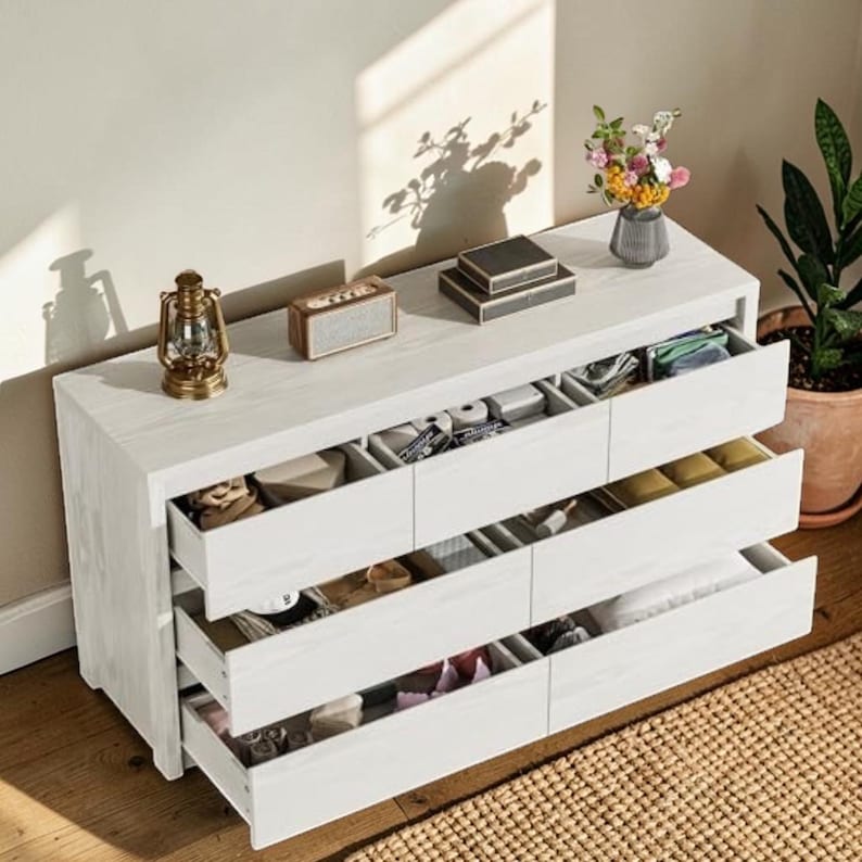 Puede incluir: Una c&oacute;moda de madera blanca con seis cajones, algunos de los cuales est&aacute;n abiertos, revelando compartimentos organizados. Un peque&ntilde;o altavoz, una linterna decorativa y un jarr&oacute;n con flores est&aacute;n encima. La c&oacute;moda est&aacute; sobre un suelo de madera.