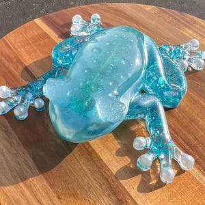 May include: A translucent blue frog figurine with a textured surface. The frog is posed on a wooden surface. The frog's body has a speckled design, and its feet are a lighter shade of blue.