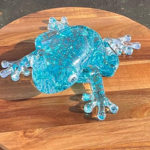 May include: A clear, turquoise frog figurine with a textured surface and visible details. The frog is positioned on a round, wooden surface. The figurine has detailed feet and a visible eye, suggesting a decorative item.