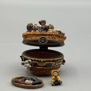May include: A detailed, open, decorative box with a boat-like design, featuring miniature animal figures and the text "S.S. Noah". The box is brown with gold accents and includes a small teddy bear figurine.