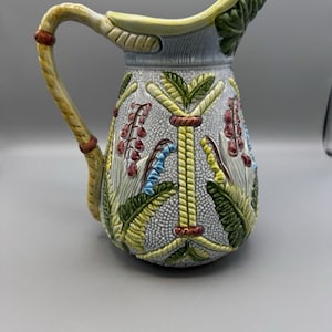 May include: A decorative ceramic pitcher with a textured, light gray background. The pitcher features a yellow handle and spout, with green, red, and blue floral and leaf designs. The handle has a rope-like design with red accents.