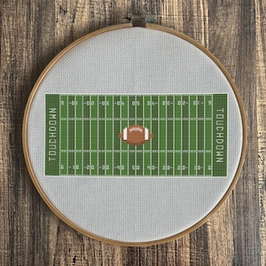 Op de afbeelding: Een ronde houten borduurring met een kruissteekpatroon van een groen American footballveld op witte stof. Het veld is voorzien van witte lijnen, cijfers en het woord TOUCHDOWN aan beide uiteinden, met een bruine football in het midden.
