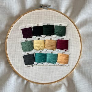 May include: A wooden embroidery hoop with white fabric and a grid pattern. Twelve spools of embroidery thread in various colours are arranged in rows. Colours include burgundy, black, dark green, and teal. The hoop is set against a white background.
