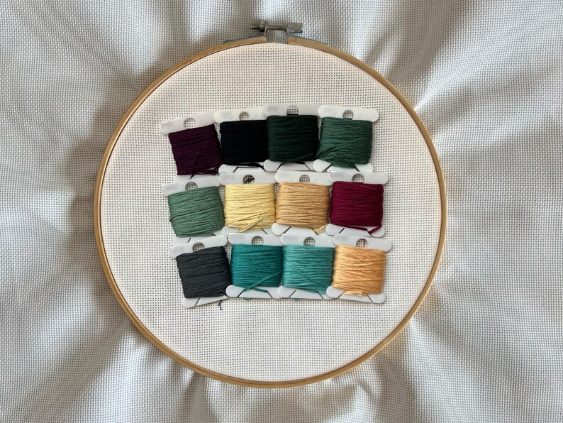 May include: A wooden embroidery hoop with white fabric and a grid pattern. Twelve spools of embroidery thread in various colours are arranged in rows. Colours include burgundy, black, dark green, and teal. The hoop is set against a white background.