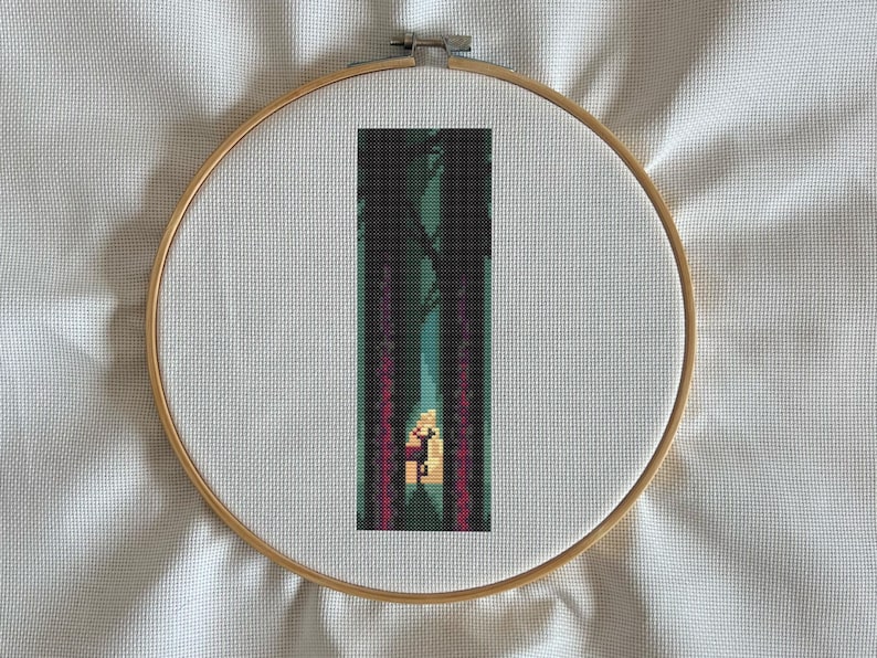 May include: Cross-stitch embroidery of a forest scene, framed in a wooden hoop. The design features a path through dark trees with a figure in the distance, rendered in shades of green, purple, and yellow.