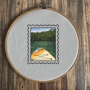May include: A cross-stitch embroidery hoop with a white fabric background. The fabric features a black and white stamp design with a green, blue, and yellow image of a kayak on a lake.