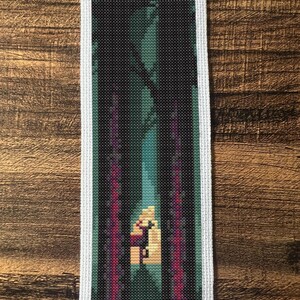 May include: A cross-stitch bookmark showing a forest scene. The design features a dark forest with tall trees, a bright opening, and vertical magenta and teal accents. The bookmark has a white border and is set against a wooden background.