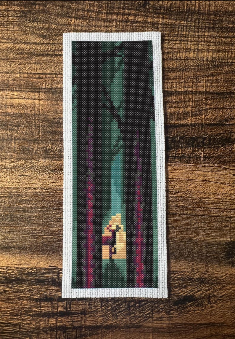 May include: A cross-stitch bookmark showing a forest scene. The design features a dark forest with tall trees, a bright opening, and vertical magenta and teal accents. The bookmark has a white border and is set against a wooden background.