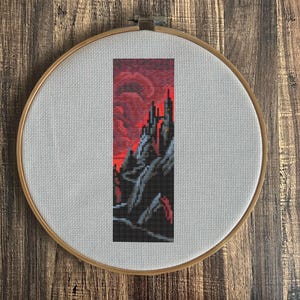 May include: A cross-stitch embroidery of a dark castle scene with a red sky, mounted in a wooden embroidery hoop. The castle is detailed in black and grey, with a fiery red sky in the background. The hoop is set against a wooden surface.