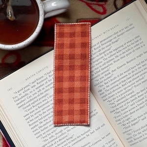 May include: A handmade bookmark with a red and orange plaid pattern, placed on an open book. The bookmark has a white border and is rectangular. A cup of tea is visible in the background.