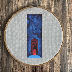 May include: Cross-stitch art in a wooden embroidery hoop. The design features a tunnel with a red arched opening, set against a blue and black background. The piece is mounted on a light gray fabric.