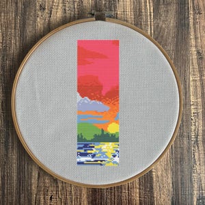 May include: A cross-stitch embroidery hoop with a colorful sunset scene. The scene features a pink sky, orange clouds, green trees, and a blue lake with white waves.