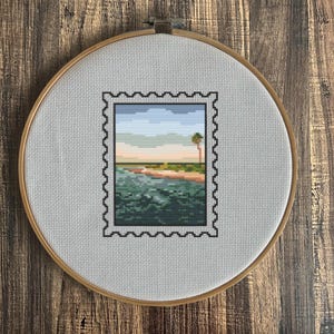 May include: Cross-stitch embroidery of a coastal landscape within a postage stamp design. The scene features a beach, ocean, and palm tree under a blue and pastel sky. The embroidery is set in a wooden hoop.