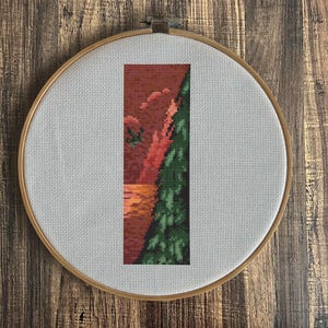 May include: A cross-stitch embroidery hoop with a landscape design. The artwork features a vertical rectangular composition with a red and orange sky, a green tree, and a textured orange foreground. The hoop is made of wood.