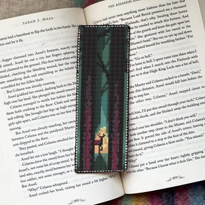 May include: A cross-stitch bookmark depicting a forest scene with a central figure. The bookmark is rectangular and rests on an open book with text by "Sarah J. Maas". The book is open to page 919.